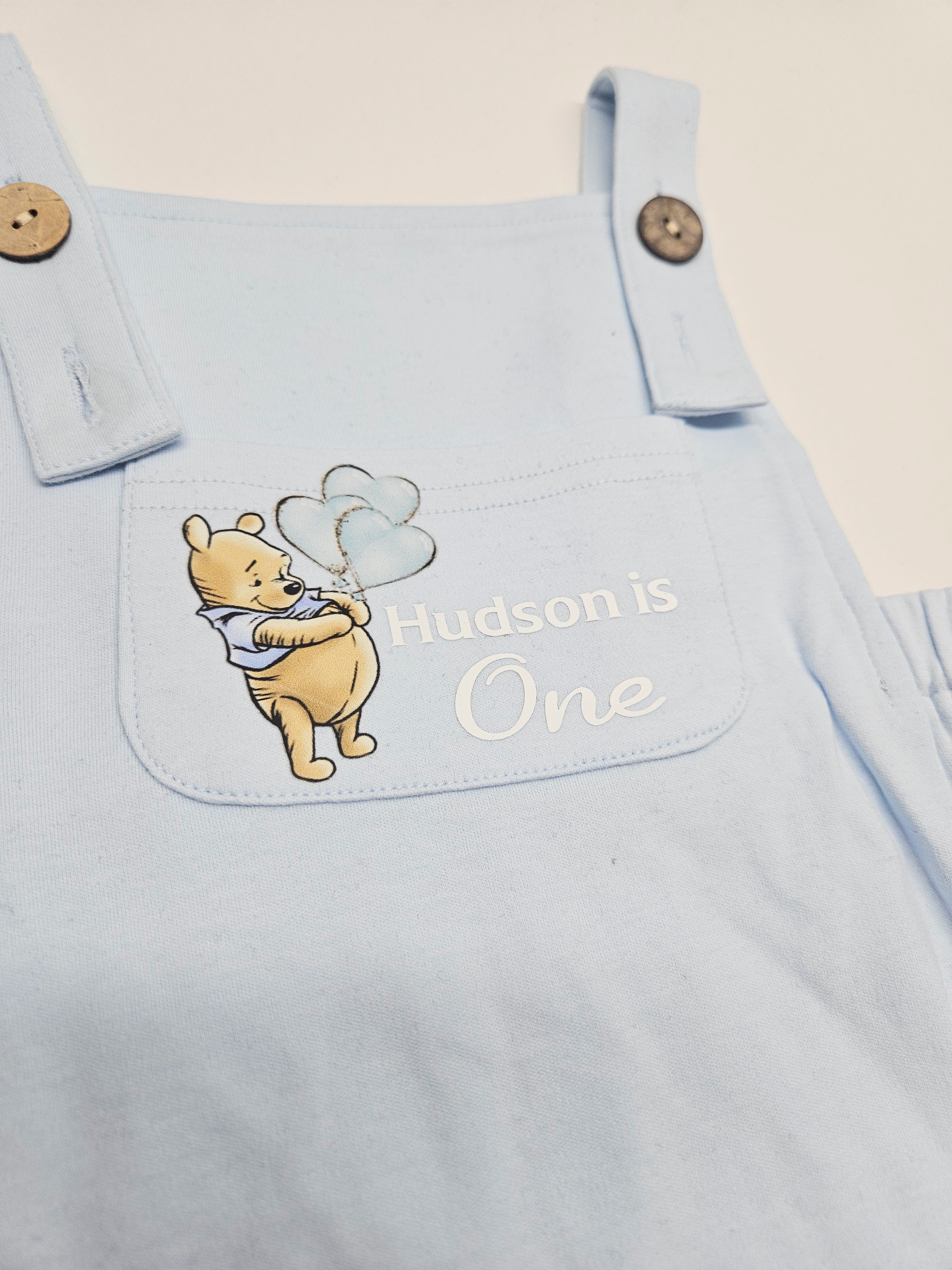 A close-up of a light blue baby overalls or romper with a front pocket featuring an embroidered Winnie the Pooh holding balloons and the text 'Hudson is One'.