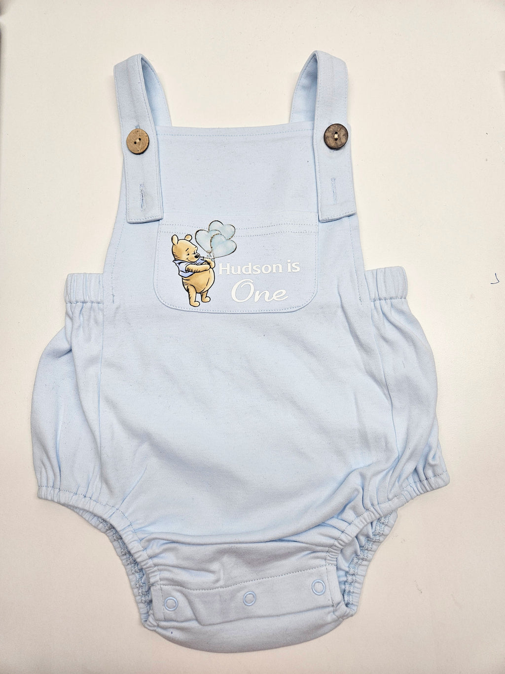 A light blue baby overalls romper with a front pocket featuring a Winnie the Pooh graphic and the text 'Aldson is One' in white script. The romper has adjustable shoulder straps with wooden buttons and a snap-button crotch.