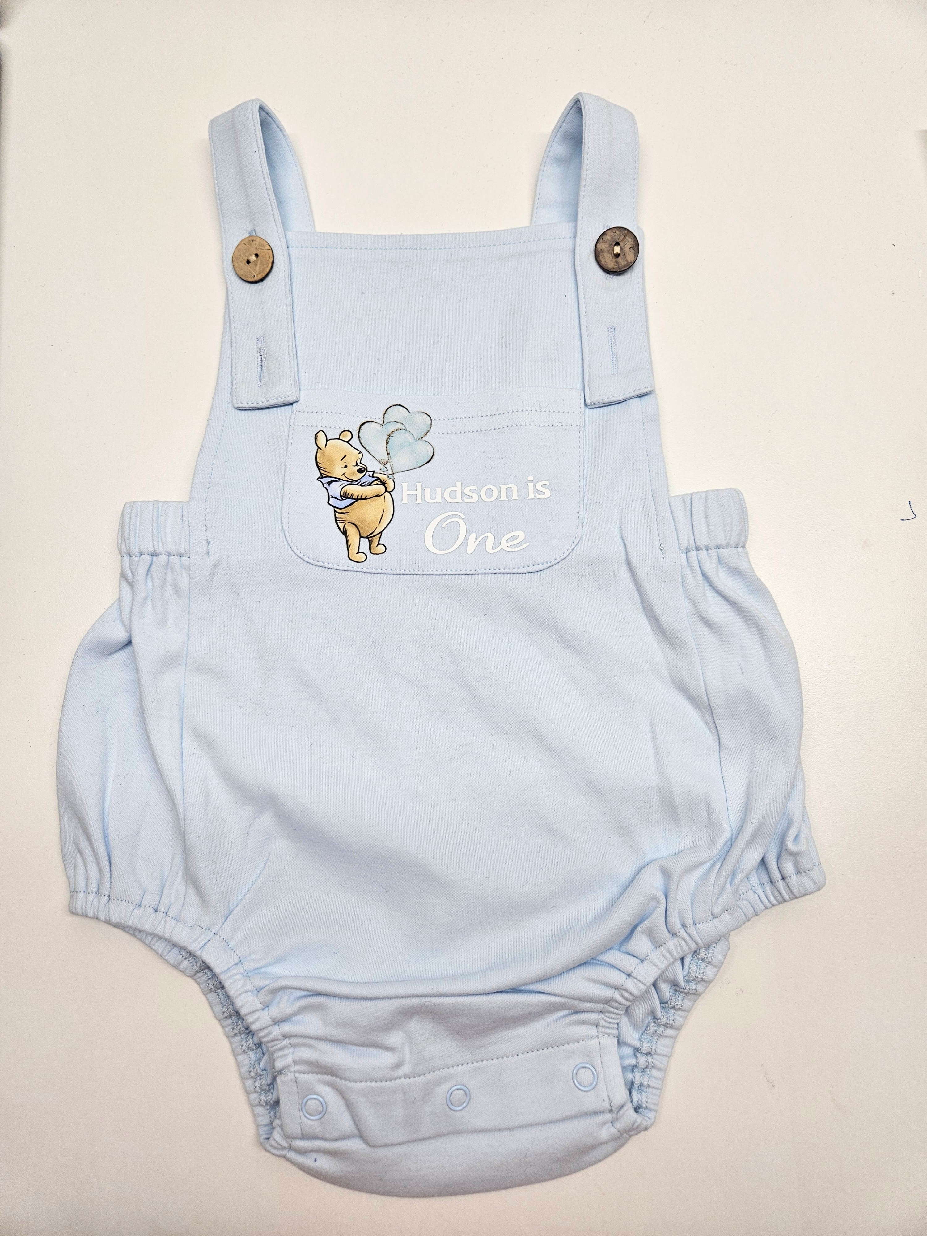 A light blue baby overalls romper with a front pocket featuring a Winnie the Pooh graphic and the text 'Aldson is One' in white script. The romper has adjustable shoulder straps with wooden buttons and a snap-button crotch.