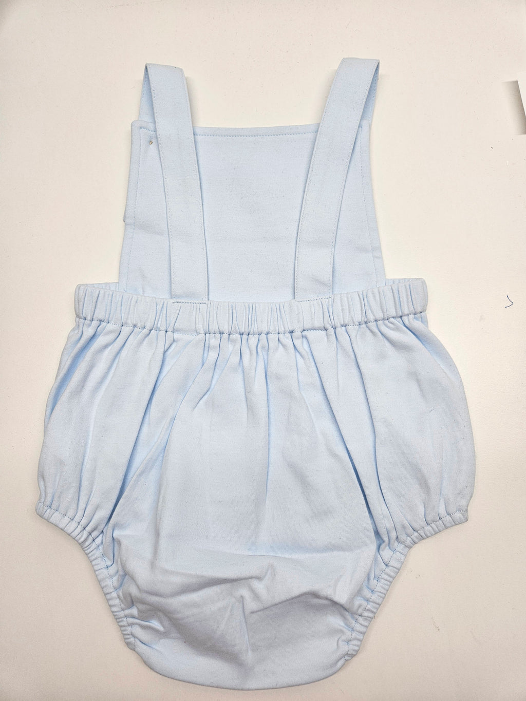 A light blue baby romper with wide shoulder straps and a gathered elastic waist, laid flat on a light beige background.