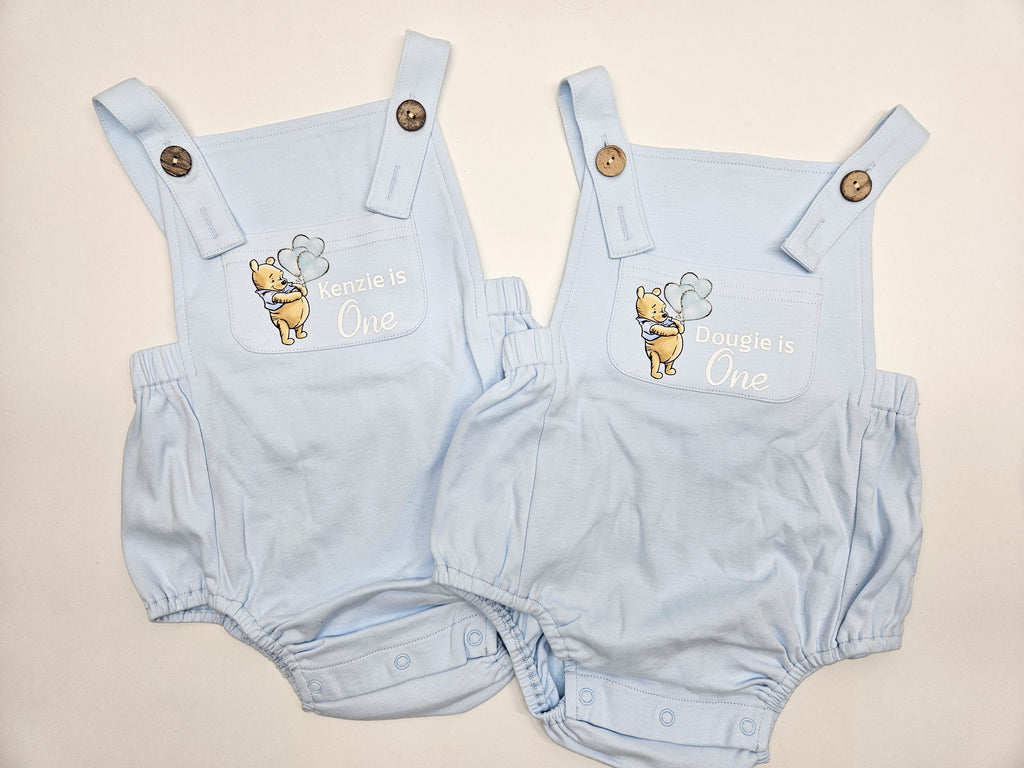 Two light blue baby bubble romper outfits laid flat on a light beige background. Each romper has a chest design featuring Winnie the Pooh and the text 'Kenzie is One' and 'Dougie is One' respectively, with adjustable shoulder straps and snap closures.