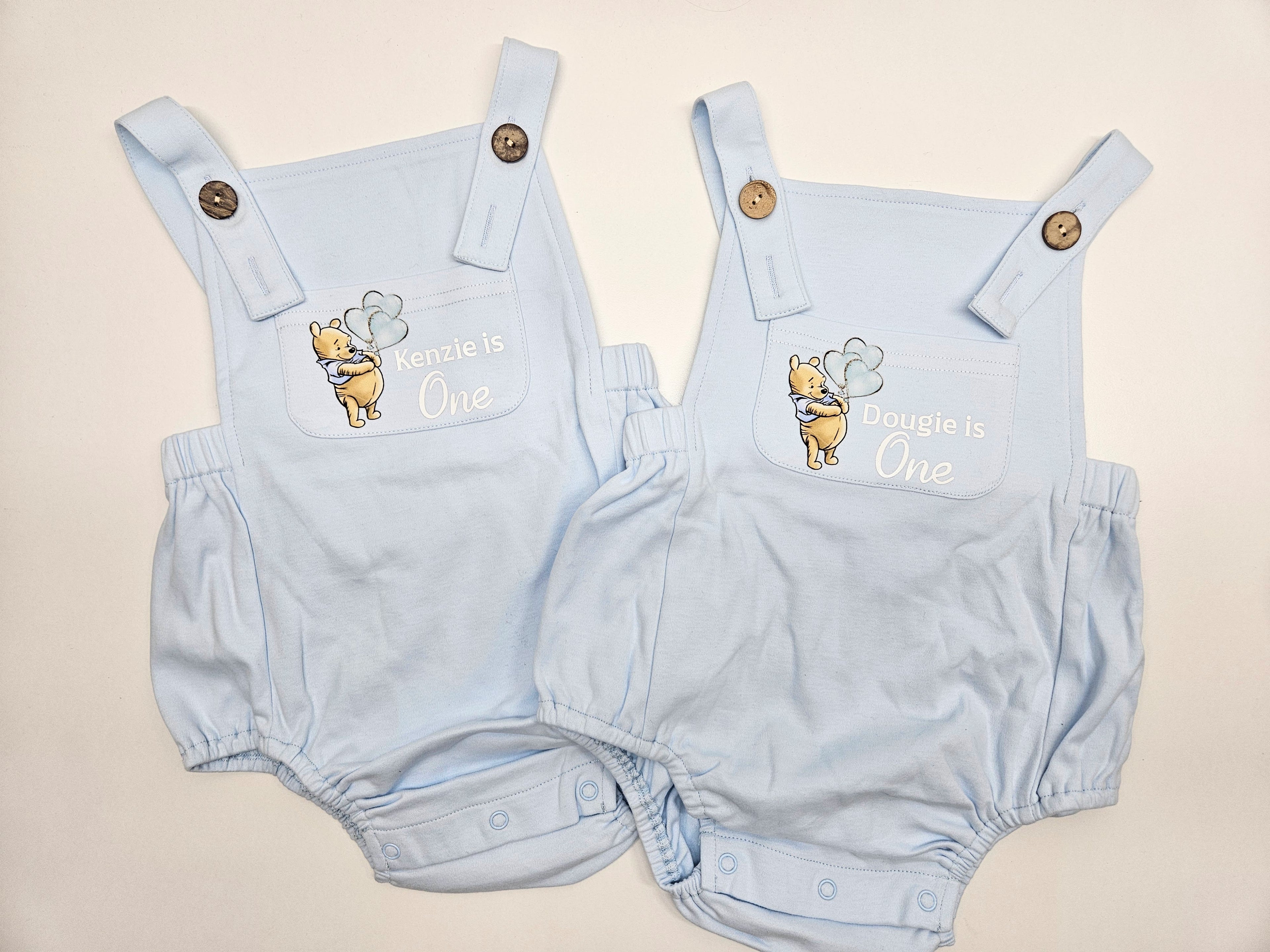 Two light blue baby bubble romper outfits laid flat on a light beige background. Each romper has a chest design featuring Winnie the Pooh and the text 'Kenzie is One' and 'Dougie is One' respectively, with adjustable shoulder straps and snap closures.