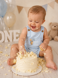 A baby wearing a light blue 'Winnie the Pooh One Year' romper sits on a wooden floor, smiling and reaching for a white frosted cake. The background features balloons, a teddy bear, and 'ONE' lettering.