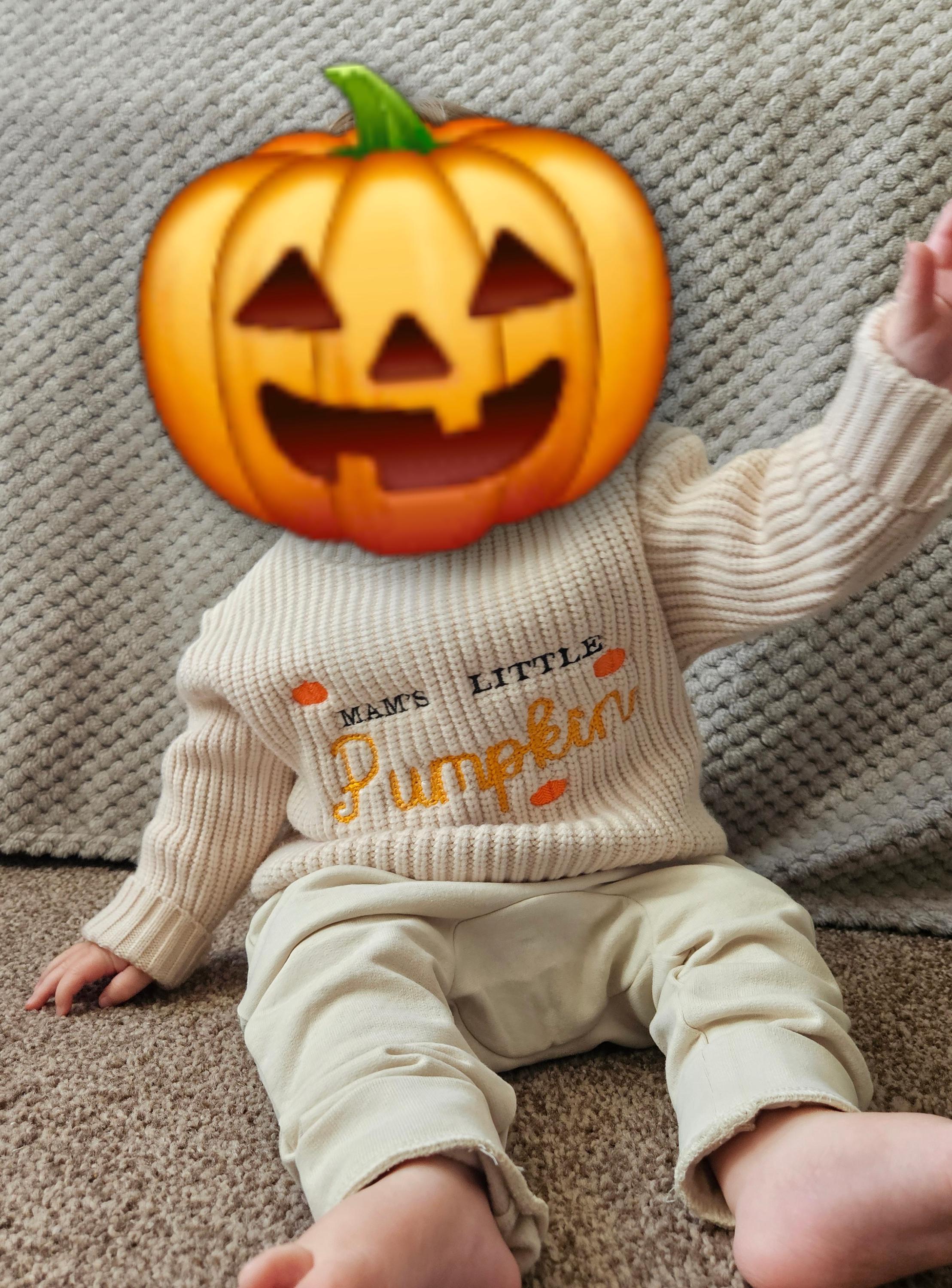 Baby wearing a sweater with 'Mama's Little Pumpkin' text, sitting on a carpeted floor.